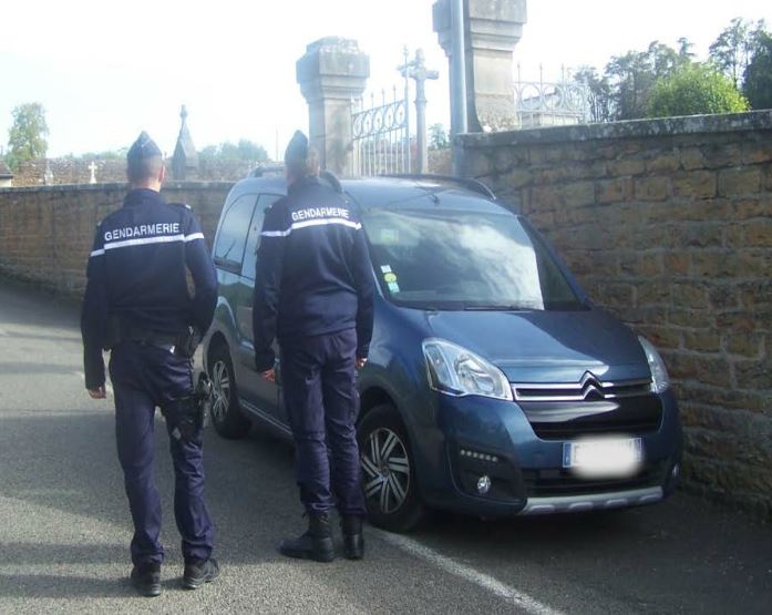 Deux gendarmes devant une voiture de gendarmerie aux abords d'un cimetière 